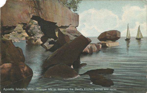 Colorized postcard of the arches and rocks comprising the Devil's Kitchen, a geological formation on Presque Isle or Stockton Island, part of the Apostle Islands. Two sailboats are on the far right. Caption reads: "Apostle Islands, Wis. Presque Isle or Stockton, the Devil's Kitchen, arches and rocks."