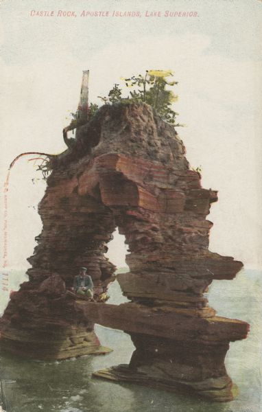 Colorized postcard of Castle Rock, a geological formation of the coast of the Apostle Islands. A man is sitting under the arch. Caption reads: "Castle Rock, Apostle Islands, Lake Superior."