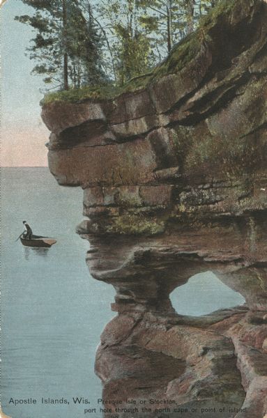 Colorized postcard of a port hole through the north cape or point of Presque Isle or Stockton Island, part of the Apostle Islands. A person in a boat is out on the lake. Caption reads: "Apostle Islands, Wis. Presque Isle or Stockton, port hole through the north cape on point of island."