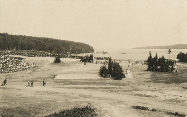 Photographic postcard of a scene from the Apostle Island Indian Pageant. A large group of young children in white shirts are sitting along a sloped hill near the shoreline. Tepees are hidden behind scattered trees, and people in Native American dress are near the shore. Parked cars are in the far background, and boats are out on the water.