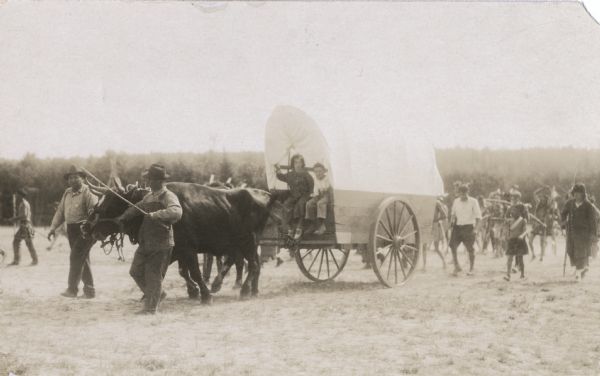 Photographic postcard of a scene from the Apostle Island Indian Pageant. Two children are sitting aboard a covered wagon pulled by a pair of steers while two men, one holding a whip, is leading the animals along. Other children and adults, some in traditional Native American costume, are following behind.