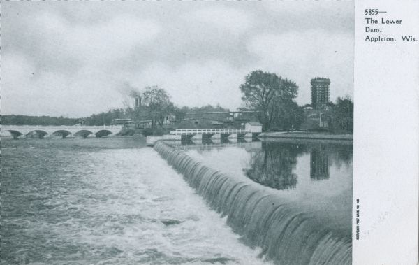 View across the lower dam and bridges near the far shoreline along the Fox River. A tall building is in the background on the right. Caption reads: "The Lower Dam, Appleton, Wis."