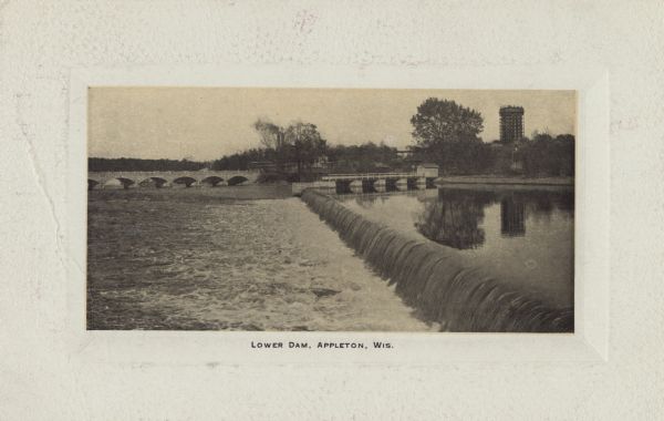 View across the lower dam on the Fox River. Bridges are near the far shoreline, and a tall building is in the distance on the right. Caption reads: "Lower Dam, Appleton, Wis."
