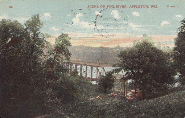 Colorized postcard looking down hill of the Fox River through the trees. A bridge is spanning the river, and piles of lumber are on the shoreline. Caption reads: "Scene on Fox River, Appleton, Wis."