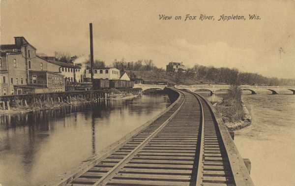 View of the Fox River near Appleton from an elevated railroad track. The fourth lock is located to the extreme right crossing the river. Railroad cars and buildings are on the left. Caption reads: "View on Fox River, Appleton, Wis."