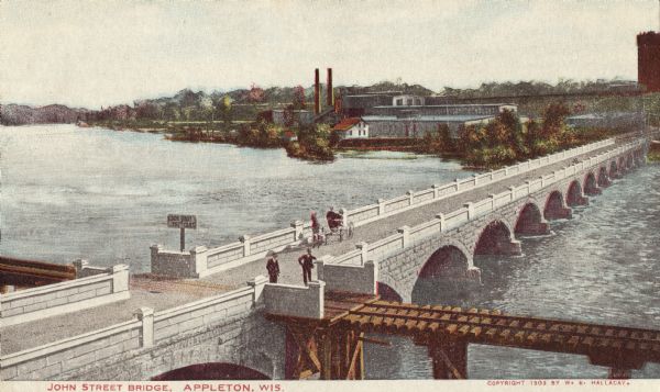Colorized postcard of an elevated view of the white brick John Street Bridge spanning the Fox River. A set of railroad tracks crosses over a section of the bridge in the foreground. Buildings and smokestacks are on the opposite shoreline. Caption reads: "John Street Bridge, Appleton, Wis."