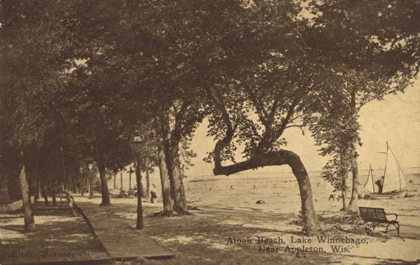 View of Aloha Beach on Lake Winnebago near Appleton. There is a shaded boardwalk along the shoreline and sailboats are on the lake. Caption reads: "Aloha Beach, Lake Winnebago, Near Appleton, Wis."