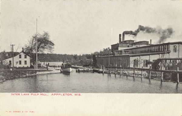 View of the Inter Lake Pulp Mill. Smoke is billowing out of the mill's smokestack. A boat is crossing the canal between two lakes. Caption reads: "Inter Lake Pulp Mill, Appleton, Wis."