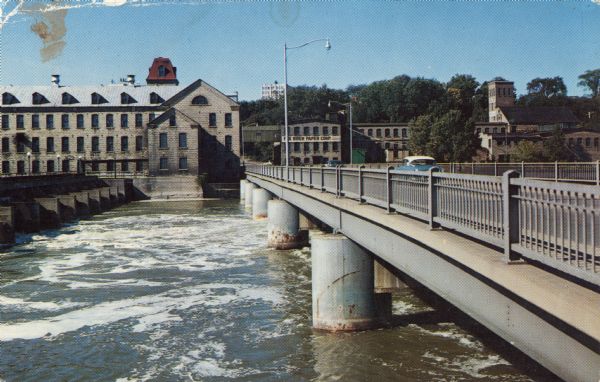 Color postcard view of the Fox River Bridge. Two cars are driving across the bridge towards the Fox River Paper Corp.
