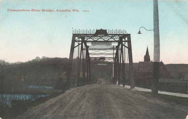 Colorized postcard of the Trempealeau River Bridge. A lamppost is in the foreground, and buildings, including a church, are in the background. Caption reads: "Trempealeau River Bridge, Arcadia, Wis."