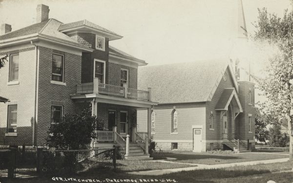 Exterior view of the German Lutheran Church and Parsonage. The one-story church with stained glass windows and steeple is in the background. The two-story brick parsonage is in the foreground. Caption reads: "Ger. Lutheran Church — Parsonage, Arcadia, Wis."