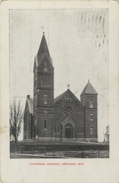 Exterior view of the front entrance to Our Lady of Perpetual Help Catholic Church. A bare tree is on the left and a fence lines the walkway to the church in the foreground. Caption reads: "Catholic Church, Arcadia, Wis."