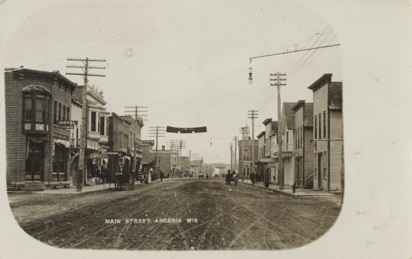 View looking down Main Street. Shops and businesses line both sides of the street including the P.J. Wossenburger Hotel, city livery, bakery and harness shop. People are walking along the sidewalks and horse-drawn wagons are along the road. There is a street light hanging over the street. Caption reads: "Main Street, Arcadia, Wis."