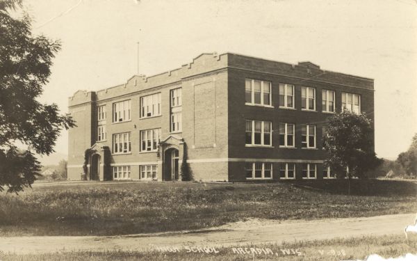 Exterior view of the two-story brick High School building with basement. A bare flagpole can be seen on the roof. Caption reads: "High School, Arcadia, Wis."