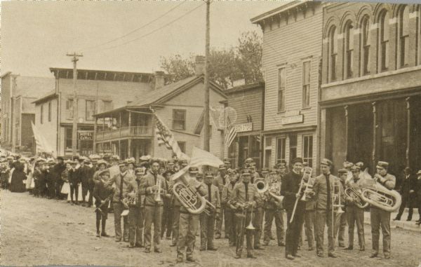 Brass marching band in uniforms and spectators at a Memorial Day parade. The band and spectators stand in the road outside city shops and business, including a bookstore and hotel. Several parade-goers hold American flags.