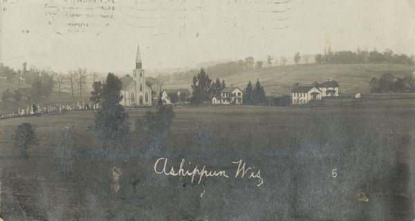 Elevated view of a church with cemetery and farmhouses and rolling hills in the distance. Caption reads: "Ashippun, Wis."