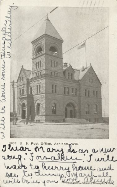 Front facade of the three-story brick U.S. Post Office with bell tower. An American flag is flying above the building. Three men are standing next to a telephone pole outside the building to the left.
