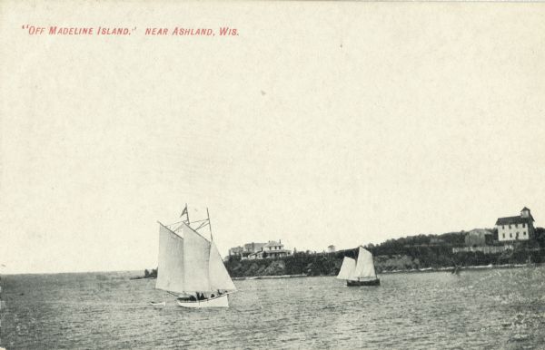 View across water towards two schooners off the shore of Madeline Island, one of the islands that comprise the Apostle Islands in Lake Superior. Two large two-story houses are on the shore in the distance.