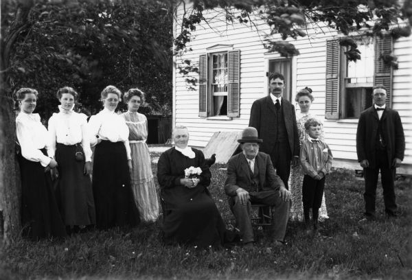 Possibly an anniversary group portrait of the couple seated in the center, with four people posed on each side of them. The women in the center holds flowers and a chicken can be seen near the house behind her. The windows of the house are open.