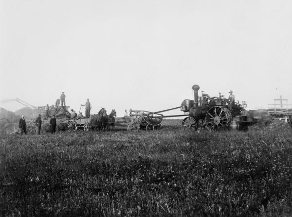 Crew of men harvesting grain using horses, a tractor and a thresher. Three men standing in the field at left are wearing suits and hats.