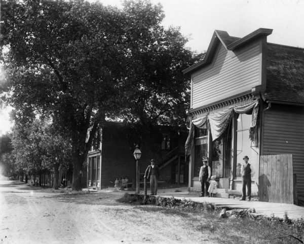 Three men and a young girl stand on a wooden sidewalk on the right in front of what appears to be a storefront. A group of four small children stand against a wall further down the street. Trees line the rest of the street in the business district. There is a street light on the nearest corner.