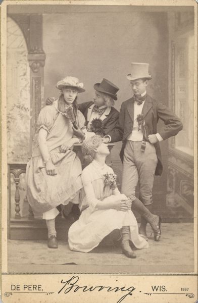 Cabinet card of a group portrait of four men in front of a painted backdrop. Two are dressed in male formal attire, with top hats. The other men are dressed in female attire, including dresses, hats, stockings and shoes. Three of the men are also wearing wigs.