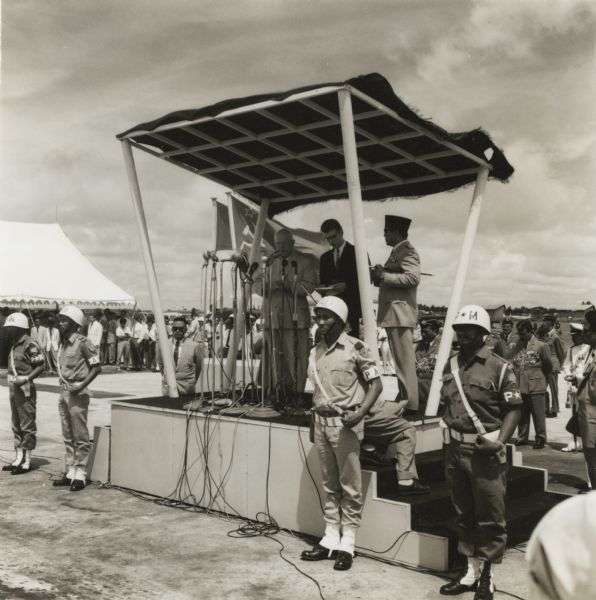 Nikita Khruschev of the Soviet Union speaks to a crowd at the airport in Indonesia. President Sukarno stands at his side, while Indonesian troops stand at attention.