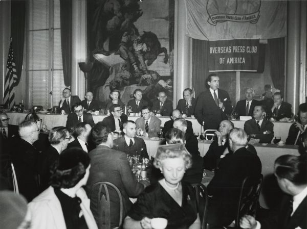 Edward R. Murrow, standing near the banner, speaks to a special Overseas Press Club luncheon at the Waldorf Astoria honoring CBS correspondents. Immediately to Murrow's left is Cecil Brown, a former CBS correspondent, who was then the president of the OPC.