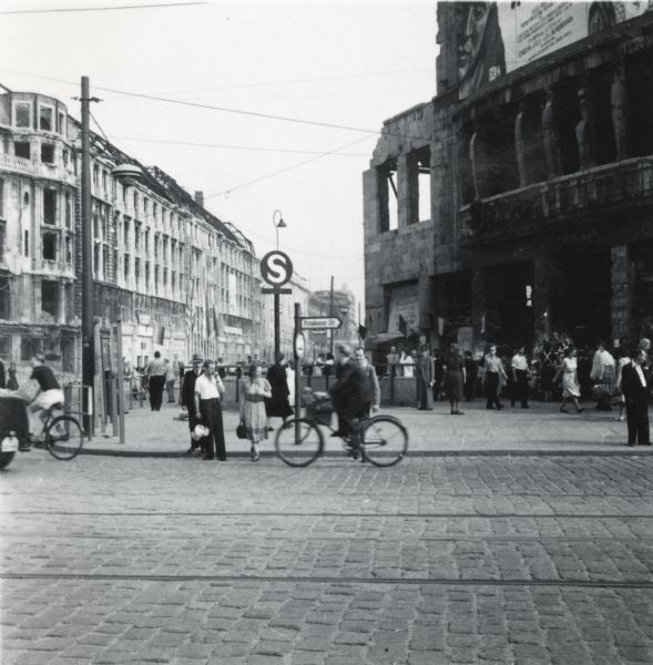A street scene in Berlin that shows the damage caused by wartime bombing. A banner or billboard on the damaged building on the right is advertising a film from DEFA.
