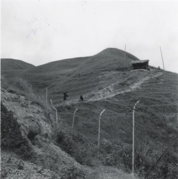 A hill in the countryside near Hong Kong, showing the fence that marked the border with Communist China. Two children are on a path on the side of the hill, and another person sits near a hut above them.