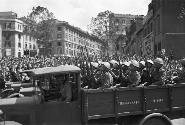 Italian gas bomb throwers, wearing their gas masks, riding in a truck during a parade for the anniversary of the founding of the Italian Empire in Rome. This parade was reviewed by King Victor Emmanuel, Benito Mussolini and Gen. Walter Von Brauchtisch, Nazi.