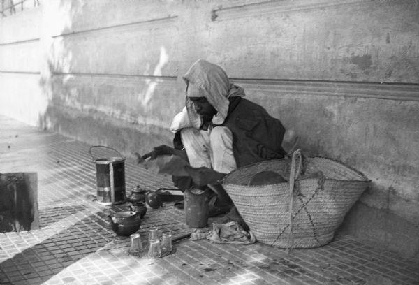 A tea seller squats with his wares on a sidewalk in Tripoli, Libya. This photograph was taken by American journalist Cecil Brown, who had a great affinity for photographing local street life.