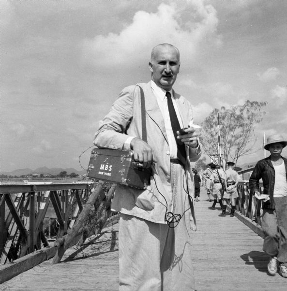 Journalist Cecil Brown, then with the Mutual Broadcasting Network, carrying the portable recorder that he used for interviewing while he was reporting on overseas news stories. He is standing on a bridge, and there are soldiers and civilians in the background.
