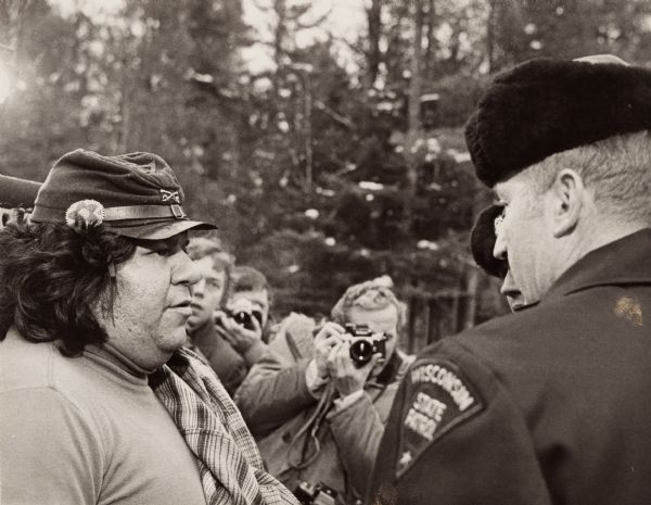 Negotiations between Neil Hawpetoss, a leader of the Menominee Warrior Society that had seized the Alexian Novitiate at Gresham in January 1975, and a member of the Wisconsin State Patrol. Eventually Governor Lucey called in the Wisconsin National Guard. While the two men talk, a photographer in the background takes their picture.