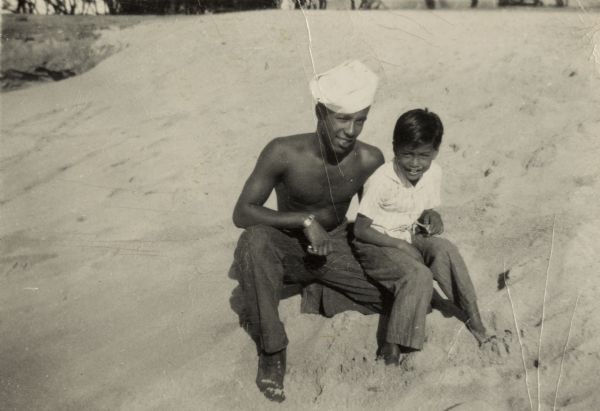 Lewis Arms poses on a beach in the Pacific Islands with a Korean boy. Arms is wearing his Navy uniform hat.