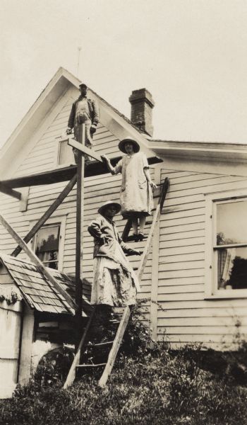 Bernard Arms is standing high on a scaffold attached to a house, while two women from the family of his neighbors, the Nofsingers, are standing below him on a ladder propped up against the house.