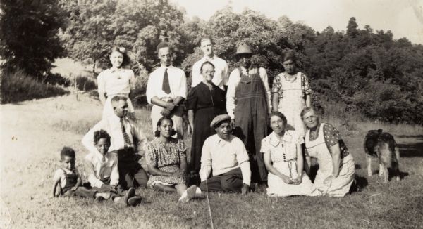 A group portrait of family and friends. From left to right, sitting in front, are Merwyn Arms, Edward Arms, Algie Shivers (kneeling), Blanche, Ed Shivers, Betsy Nofsinger, and Mert Nofsinger. Standing, from left to right are Eloise Arms, Otis Arms, Florie (in black dress), Raymond Schmitt (standing behind Florie), Tom Shivers, and Mrs. B. There is a dog standing on the far right.