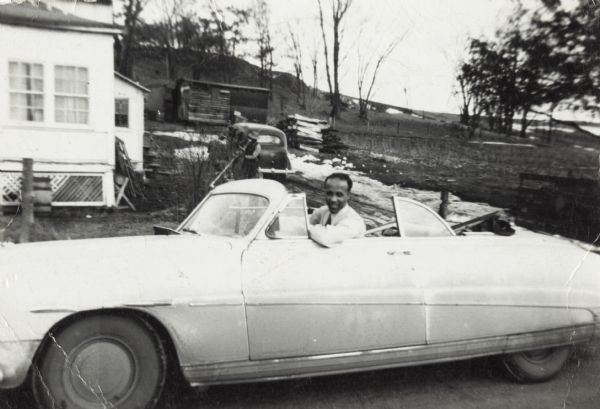 Lewis Arms is seated in a 1949 yellow Hudson convertible at Bernard Arms's farm. A man is standing in the background near another automobile parked near the farmhouse.