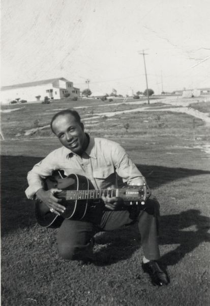 Lewis Arms kneels close to the ground while playing a guitar. There is a large, industrial looking building in the far background.