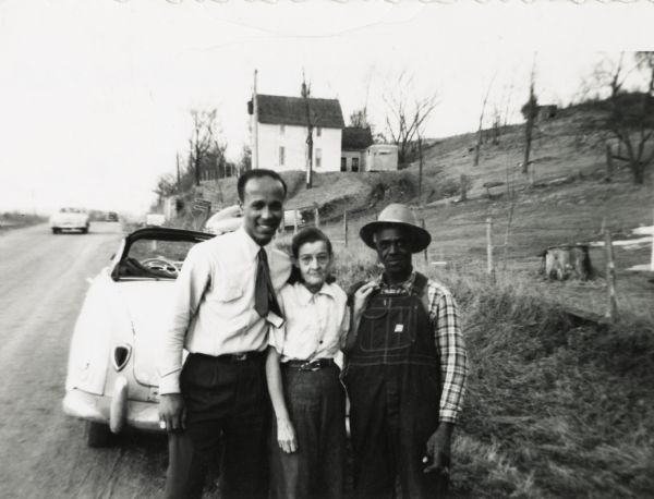 From left to right are Lewis, Nellie and Bernard Arms posing behind a convertible on Old Lake Road, with Bernard and Nellie's house in the background.