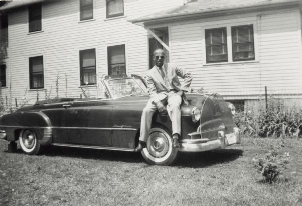 Lewis Arms, dressed in a suit and sunglasses, sits on the hood of his 1949 Pontiac automobile.