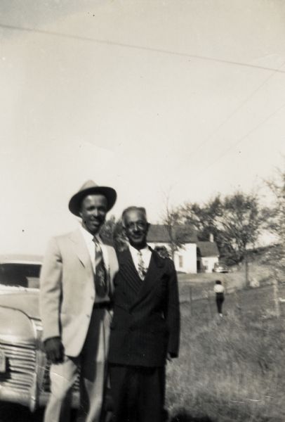 Lewis Arms (left, wearing a hat) poses with his arm around his uncle Otis Arms, probably at Bernard Arms's farm. There is a woman in a field walking away in the background.