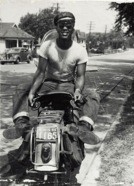 Lewis "Honeylove" Arms is seated backwards on his 1949 Harley-Davidson motorcycle. He is wearing a hat and sunglasses.