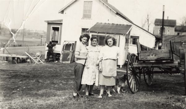 Posing together from left to right are Elinora, Nellie Arms (aunt of Lewis Arms), and Bonnie Childs. They are at Nellie and Bernard Arms's farm. There is a dog near the wagon, and a man is working near a window of the farmhouse in the background.