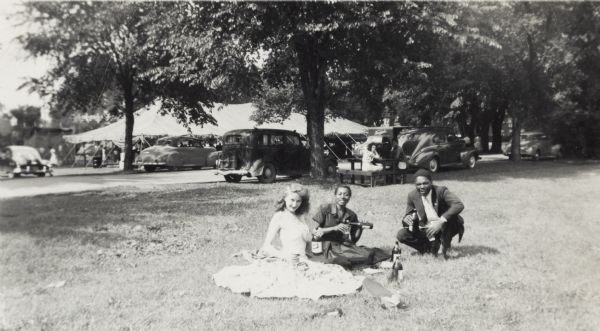 Friends of Lewis Arms enjoy beer at a party at Brittingham Park. Seated on the ground from left to right are: Elinora (mother of Lewis Arms, Jr.), Hattie, and "Boots". In the background is a festival tent and parked cars.