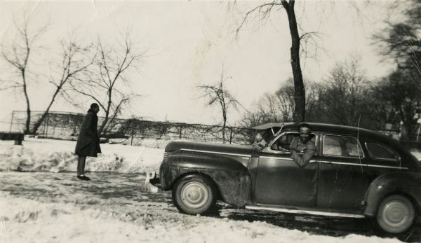 Lewis Arms is sitting in the driver's seat of a 1941(?) DeSoto automobile owned by Joe Brown, as his friend Booker T (standing) looks on. They are at James Madison Park in the winter.
