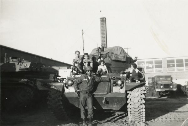 Lewis Arms poses in front of a military tank wearing army gear. On the tank from left to right are Lew Arms, Jr., Craig Arms, Rita Arms, and Mamie Arms.