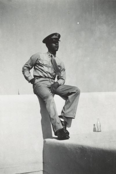 Lewis Arms sits on a short concrete wall wearing his military uniform. There is an empty soda bottle at his feet.