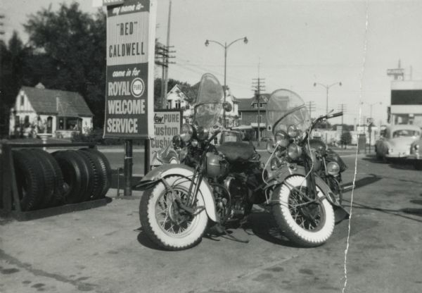 Two Harley-Davidson motorcycles belonging to Lewis Arms and Charlie Caldwell are parked on the corner of Olin Avenue and Park Street at a gas/service station.