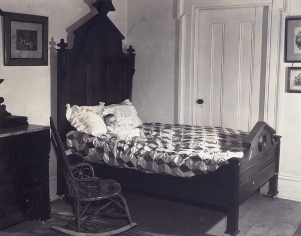 View of a bedroom in the restored Charles A. Grignon House in Kaukauna. The bed has a tall Victorian headboard and is covered with a quilt. A toddler is posed on the bed.